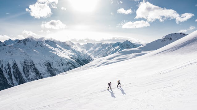 兩位登山者正沿著積雪覆蓋的山坡前行，遠方是壯闊的群山與陽光，象徵持續前行與突破困境的力量。"