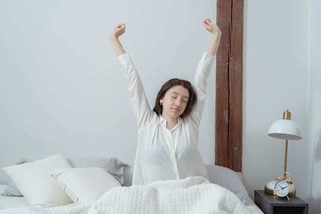 一位女性早晨在房間伸懶腰,穿著白色睡衣坐在床上,享受清晨時光