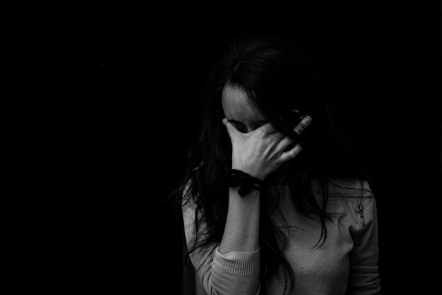 一位女性低頭掩面，身處黑暗背景中，象徵母職倦怠與自我認同的掙扎