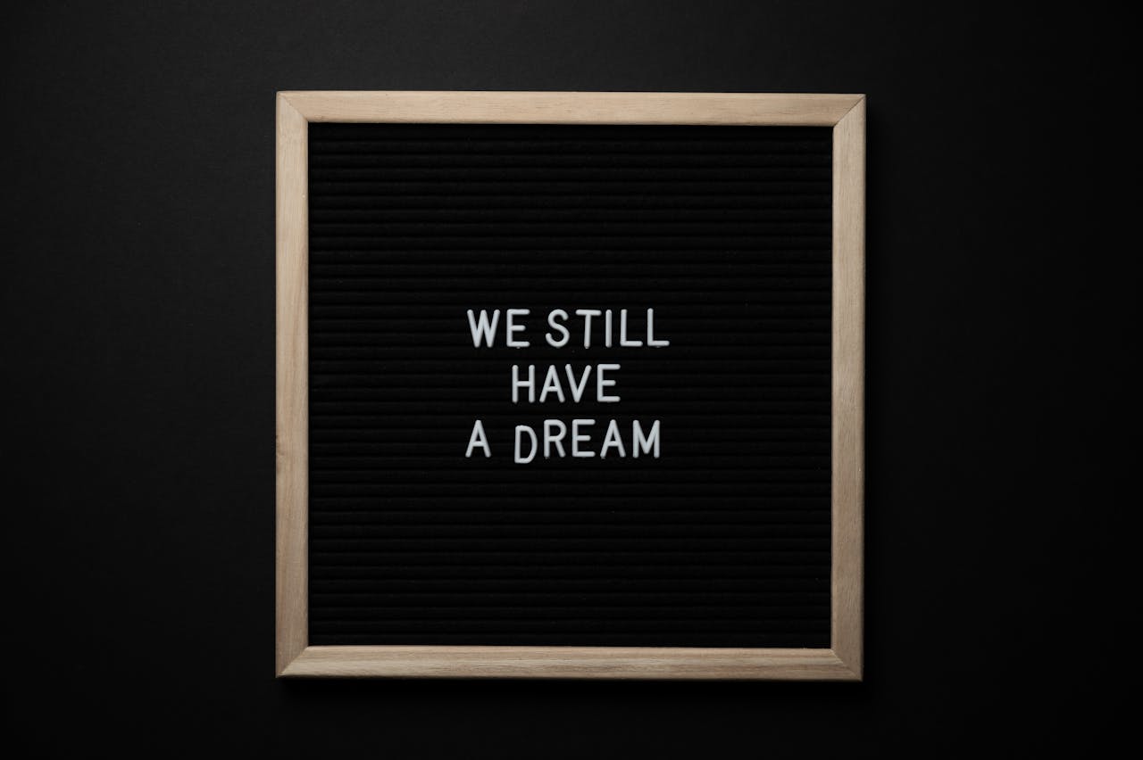 一塊黑色佈告板上，以白色字母拼出「WE STILL HAVE A DREAM」，背景沈穩，象徵未熄的夢想。