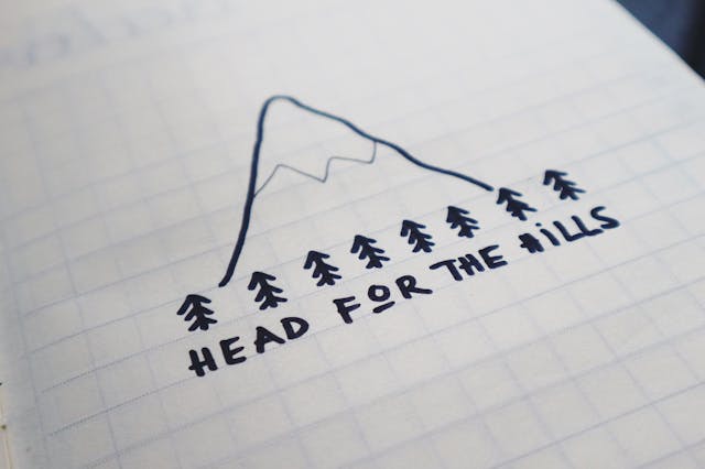 手繪山峰與松樹插畫，底下寫著「HEAD FOR THE HILLS」的字樣，畫在方格紙上。
