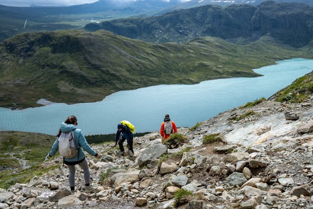 三位登山客沿著岩石小徑下山，背後是湛藍的湖泊與壯麗山景