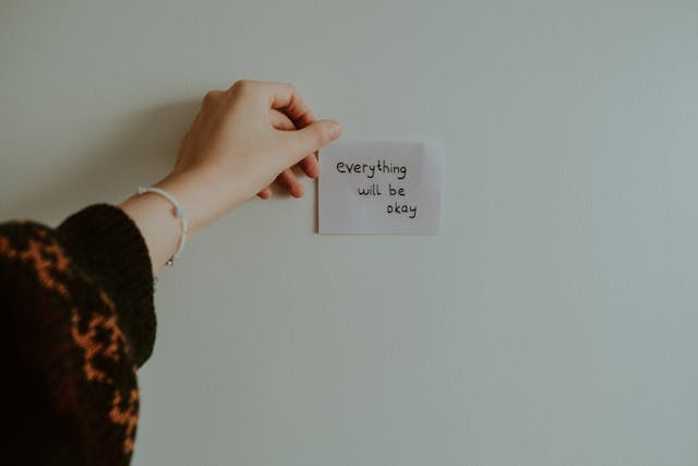 手持自我肯定紙條，上面寫著「everything will be okay」，背景簡約白牆。