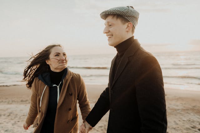 一對情侶在夕陽下的海邊散步,牽著手微笑對望,海風吹起她的頭髮,氣氛溫柔又自在。