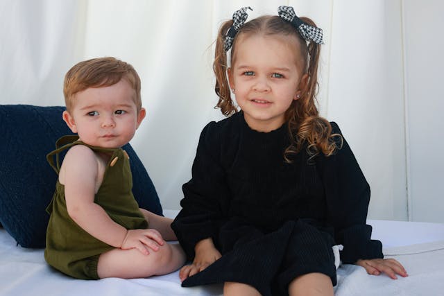一對姊弟坐在白色背景前，姊姊穿著黑色洋裝、弟弟穿著綠色吊帶褲，象徵長女與弟弟之間的童年連結與手足關係。